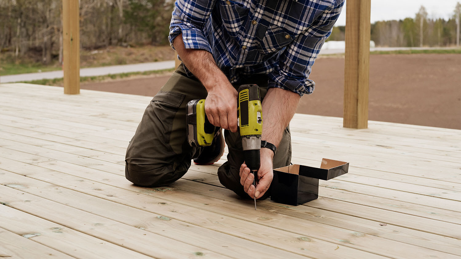 A man kneeling on a wooden deck driving a screw with a cordless drill